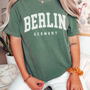 Pode incluir: Uma camiseta verde com a palavra "BERLIN" em texto branco e "GERMANY" em texto branco menor abaixo. A camiseta est&aacute; sendo usada por uma pessoa com cabelo loiro comprido e cal&ccedil;as brancas.