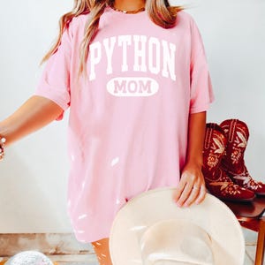 May include: A pale pink t-shirt with the white text "PYTHON MOM". The image also shows a cream-coloured hat and red cowboy boots with floral embroidery. The style is casual and feminine. The boots are on a wooden chair.