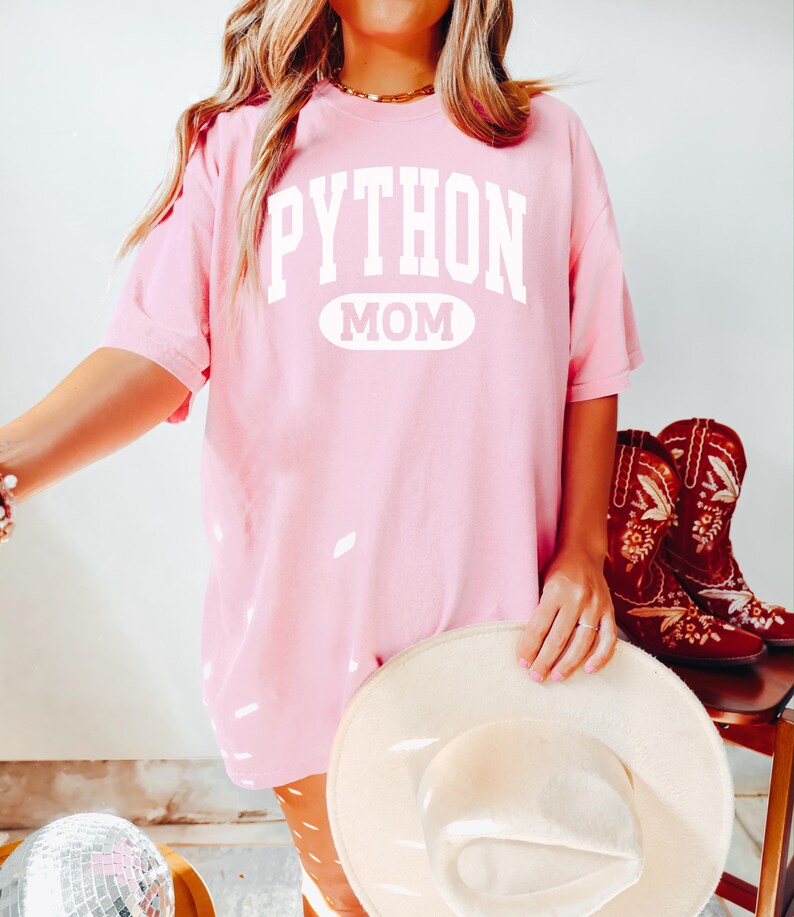 May include: A pale pink t-shirt with the white text "PYTHON MOM". The image also shows a cream-coloured hat and red cowboy boots with floral embroidery. The style is casual and feminine. The boots are on a wooden chair.