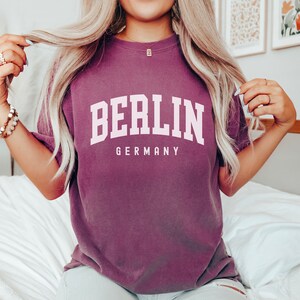 Pode incluir: Uma camiseta roxa com o texto "BERLIM ALEMANHA" em letras brancas.