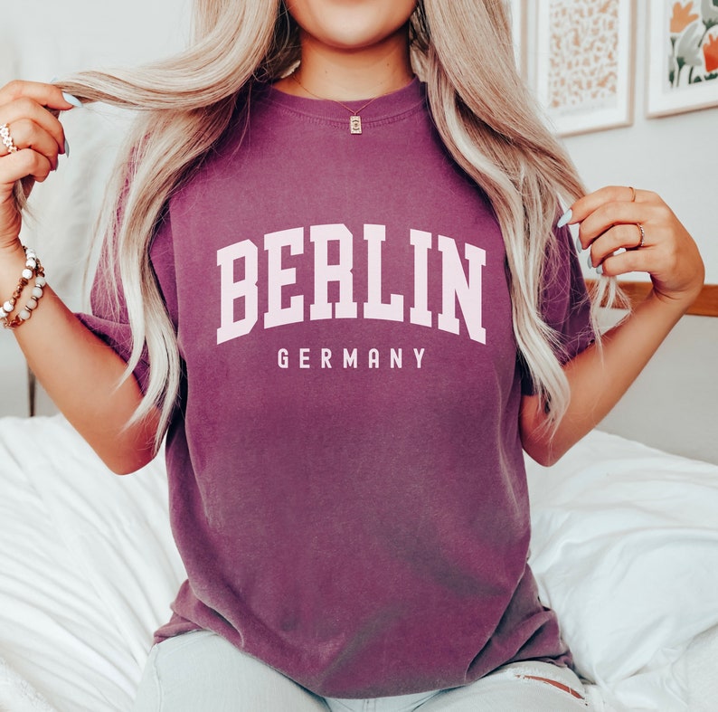 Pode incluir: Uma camiseta roxa com o texto "BERLIM ALEMANHA" em letras brancas.