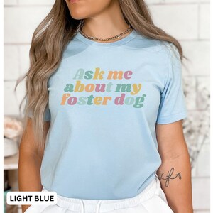 May include: Light blue t-shirt with the text "Ask me about my foster dog" in colorful retro-style lettering.