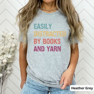 May include: Heather grey t-shirt with the text "EASILY DISTRACTED BY BOOKS AND YARN" in a retro font with colorful accents.