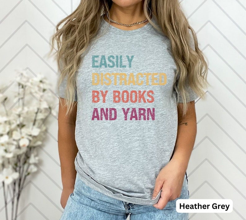 May include: Heather grey t-shirt with the text "EASILY DISTRACTED BY BOOKS AND YARN" in a retro font with colorful accents.