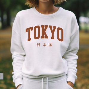 May include: White sweatshirt with a brown graphic print of the word "TOKYO" in a collegiate font with the Japanese characters for "Japan" underneath.