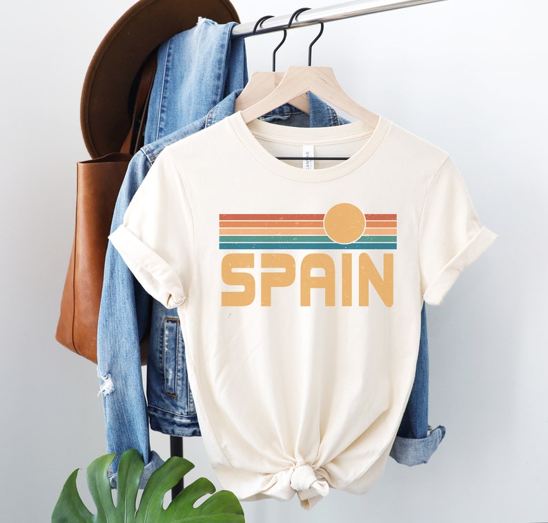 May include: A cream colored t-shirt with a retro-style graphic design featuring a sunset with orange, red, yellow, green, and blue stripes and the word "SPAIN" in a bold, yellow font.