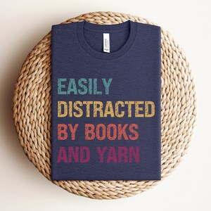 May include: A navy blue t-shirt with the text "EASILY DISTRACTED BY BOOKS AND YARN" in a vintage style font with colorful accents.
