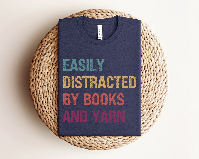 May include: A navy blue t-shirt with the text "EASILY DISTRACTED BY BOOKS AND YARN" in a vintage style font with colorful accents.