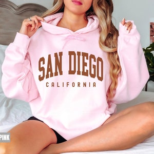Pode incluir: Um moletom rosa claro com o texto "San Diego California" em letras marrons.