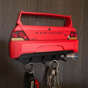 May include: A red and black key holder designed as a car's rear end, with the word "EVOLUTION" displayed. It features key hooks and an exhaust pipe detail. Keys and a car key are suspended from the hooks. The key holder is mounted on a wall.