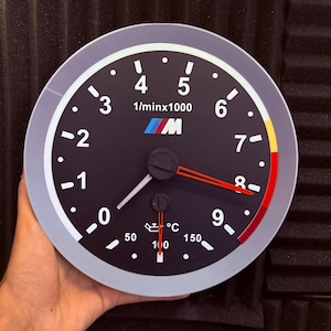 May include: A circular wall clock designed to resemble a car tachometer with a black face, white numerals from 0 to 9, and the M logo in blue, purple, and red. It features a red hour hand, a white minute hand, and a smaller oil temperature gauge.