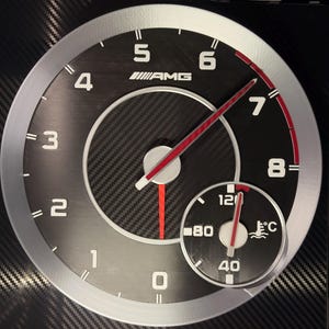 May include: A car dashboard gauge with a carbon fibre background. The main gauge has numbers from 0 to 8, with a red needle. A smaller gauge shows the temperature in Celsius. The gauge has the AMG logo.