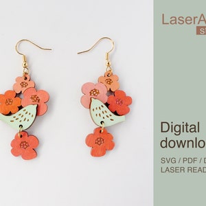 May include: A pair of laser cut wooden earrings featuring a small green bird with pink and orange flowers. The earrings are labeled "Digital download SVG/PDF/DXF Laser Ready Files" and "LaserArtist studio".