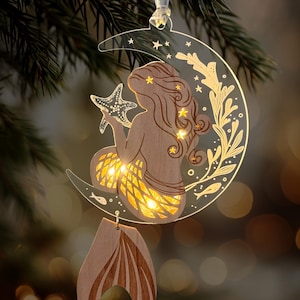 May include: A wooden mermaid ornament holding a starfish, seated on a crescent moon with star and floral accents. The mermaid's tail and the moon are illuminated with warm lights. A wooden tail hangs below, suspended by a silver ribbon.
