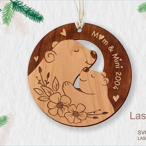 Puede incluir: Adorno navideño de madera con dos osos, uno más grande que el otro, abrazándose. El adorno está decorado con diseños florales y el texto "Mom & Mini 2004".