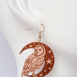 May include: Wooden earrings featuring a crescent moon with stars and a barn owl. The owl is carved in a light brown color and the moon and stars are a darker brown.