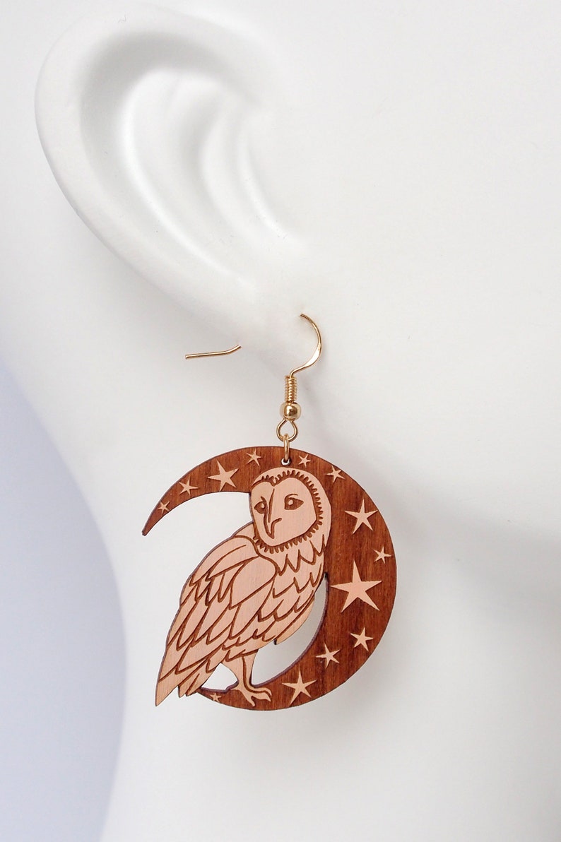 May include: Wooden earrings featuring a crescent moon with stars and a barn owl. The owl is carved in a light brown color and the moon and stars are a darker brown.