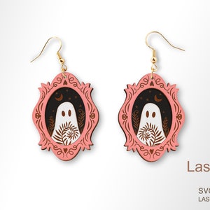Op de afbeelding: Een paar oorbellen met Halloween-thema. Elke oorbel heeft een roze, sierlijk frame met een zwart interieur. Binnenin het frame bevindt zich een wit spook met een botanisch ontwerp. De oorbellen hebben goudkleurige haken. Het LaserArtist studio-logo is zichtbaar.