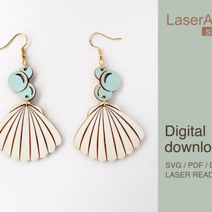 May include: A pair of laser-cut wooden earrings featuring a seashell design with a light blue circle accent. The earrings are gold-toned and have a hook closure. The text "LaserArtist studio" and "Digital download SVG / PDF / DXF LASER READY FILES" is visible in the image.