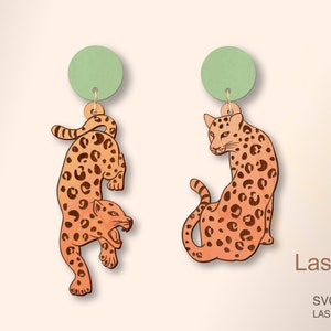 May include: A pair of laser cut wooden earrings featuring a leopard design. The earrings are orange and brown with black spots. The earrings are attached to a green circle.