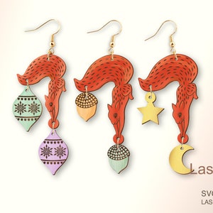 May include: Three pairs of laser-cut wooden earrings featuring a red squirrel holding a different ornament. The ornaments are a green and white snowflake ornament, a brown acorn, and a gold crescent moon.
