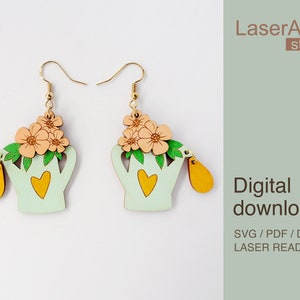 Könnte beinhalten: Ein Paar lasergeschnittene Ohrringe aus Holz mit einer Gießkanne mit Blumen. Die Gießkanne ist hellgrün mit einem gelben Herzen und einem gelben Tropfen, der am Ausguss hängt. Die Blumen sind pfirsichfarben und rosa mit grünen Blättern. Die Ohrringe befinden sich auf einem weißen Hintergrund mit dem Text "LaserArtist studio" und "Digitaler Download SVG / PDF / DXF LASER-READY-DATEIEN" in brauner Schrift.