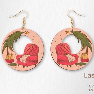 May include: Wooden hoop earrings with a festive design. Each earring features a cat sleeping in a red armchair, a Christmas tree, and gifts. The earrings have gold-coloured hooks and a light pink background.