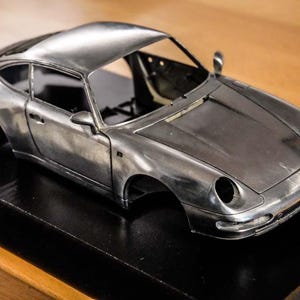 Peut inclure: Une maquette de voiture argentée, probablement une Porsche, est présentée sur une base noire. La voiture présente des phares détaillés, des rétroviseurs latéraux et une ligne de toit profilée. Le modèle est exposé sur une surface en bois.