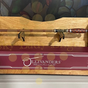 May include: A wooden wand display stand with a brown wand resting on the top. The stand has a burgundy box with the text "Ollivanders" Makers of Fine Wands since 382 BC" on the front.