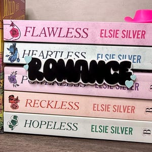 Puede incluir: Una pila de novelas rom&aacute;nticas de Elsie Silver, con t&iacute;tulos como "Flawless" y "Heartless". Un sombrero de vaquero marr&oacute;n y un sombrero de vaquero rosa est&aacute;n colocados cerca de los libros. La palabra "Romance" est&aacute; en cursiva negra.
