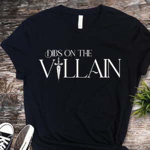 May include: Black t-shirt with the text "Dibs on the Villain" in white, featuring a sword graphic. The shirt is displayed on a wooden surface, with a small green plant and a pair of black and white sneakers visible.