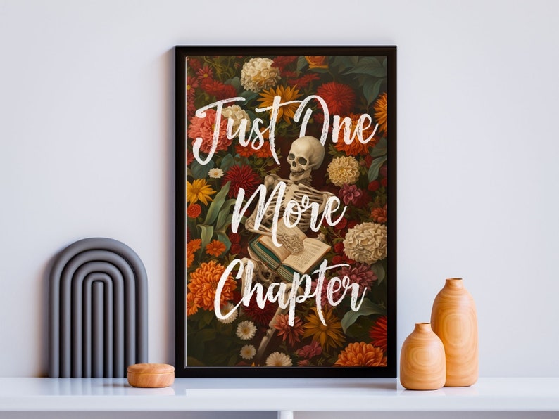 May include: A black framed print with a skeleton reading a book surrounded by colorful flowers. The text "Just One More Chapter" is written in white on the print.