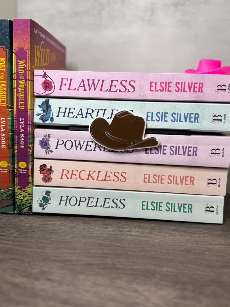 Puede incluir: Una pila de libros con t&iacute;tulos como "Flawless" y "Hopeless" de Elsie Silver. Un pin de sombrero de vaquero marr&oacute;n descansa sobre los libros. Los libros son de colores pastel, incluyendo rosa, azul y melocot&oacute;n.