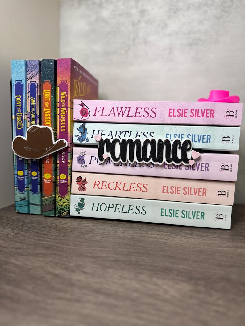 Puede incluir: Una pila de libros con t&iacute;tulos como "Flawless", "Heartless" y "Reckless" de Elsie Silver. Un marcap&aacute;ginas con forma de sombrero de vaquero marr&oacute;n y un sombrero de vaquero rosa est&aacute;n sobre los libros. La palabra "romance" es un recorte negro sobre los libros.