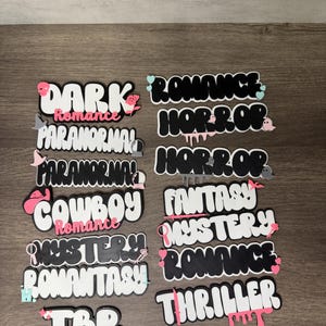 Puede incluir: Una colecci&oacute;n de piezas de arte de palabras troqueladas en blanco, negro y rosa. Las palabras incluyen "DARK ROMANCE", "HORROR", "COWBOY ROMANCE", "MYSTERY ROMANCE", "TBR" y m&aacute;s. Cada pieza tiene un dise&ntilde;o &uacute;nico con elementos decorativos.