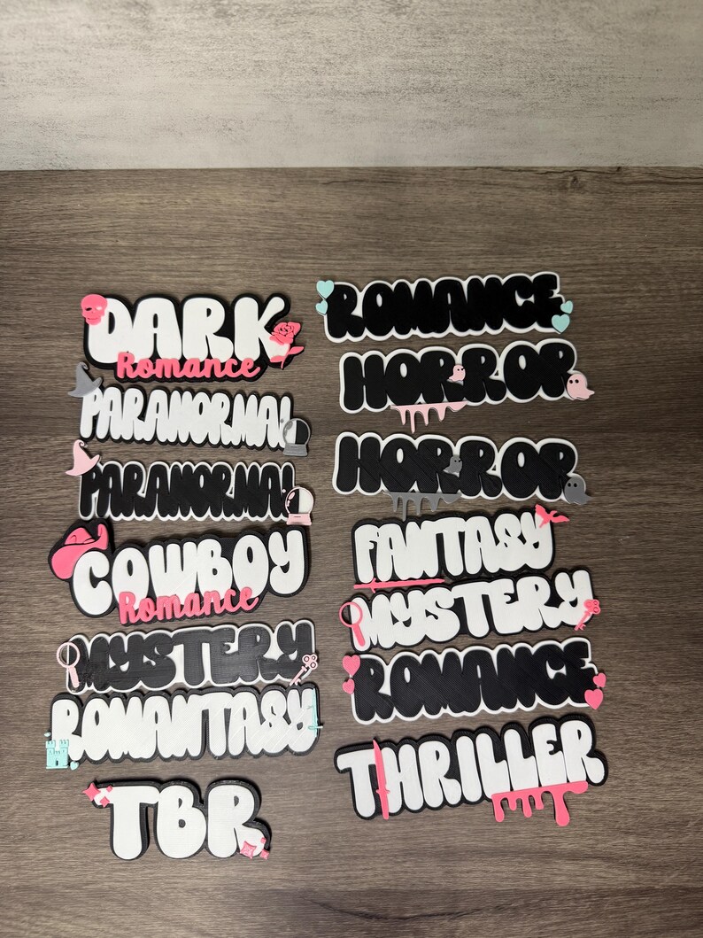 Puede incluir: Una colecci&oacute;n de piezas de arte de palabras troqueladas en blanco, negro y rosa. Las palabras incluyen "DARK ROMANCE", "HORROR", "COWBOY ROMANCE", "MYSTERY ROMANCE", "TBR" y m&aacute;s. Cada pieza tiene un dise&ntilde;o &uacute;nico con elementos decorativos.