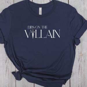 May include: Navy blue t-shirt with the text "Dibs on the Villain" in white, featuring a sword graphic. The shirt is made of soft material and has rolled-up sleeves.