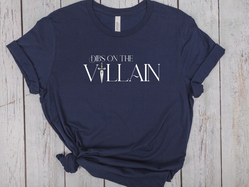 May include: Navy blue t-shirt with the text "Dibs on the Villain" in white, featuring a sword graphic. The shirt is made of soft material and has rolled-up sleeves.