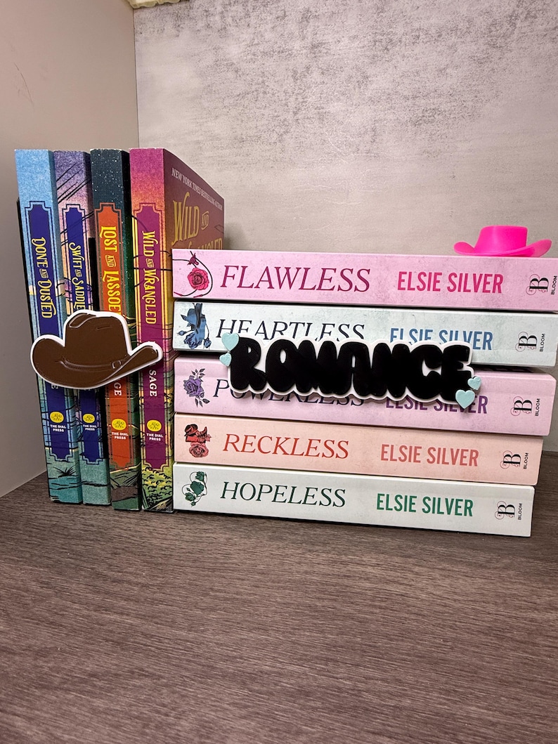 Puede incluir: Una pila de novelas rom&aacute;nticas de Elsie Silver, con t&iacute;tulos como "Flawless" y "Heartless". Un sombrero de vaquero marr&oacute;n y un sombrero de vaquero rosa est&aacute;n colocados cerca de los libros. La palabra "Romance" est&aacute; en cursiva negra.