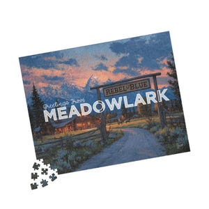 Könnte beinhalten: Fertiggestelltes Puzzle mit einer Landschaft, auf dem eine Straße zu einem Schild mit der Aufschrift „REBEL BLUE MEADOWLARK“ führt. Das Puzzle enthält den Text „Greetings From“ und eine Bergkette. Der Himmel ist eine Mischung aus Orange und Blau.