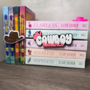 Puede incluir: Una pila de libros con cubiertas rosas, blancas y azules. Los libros se titulan "Flawless", "Reckless" y "Hopeless" de Elsie Silver. Un sombrero de vaquero marr&oacute;n y la palabra "Cowboy Romance" son visibles.