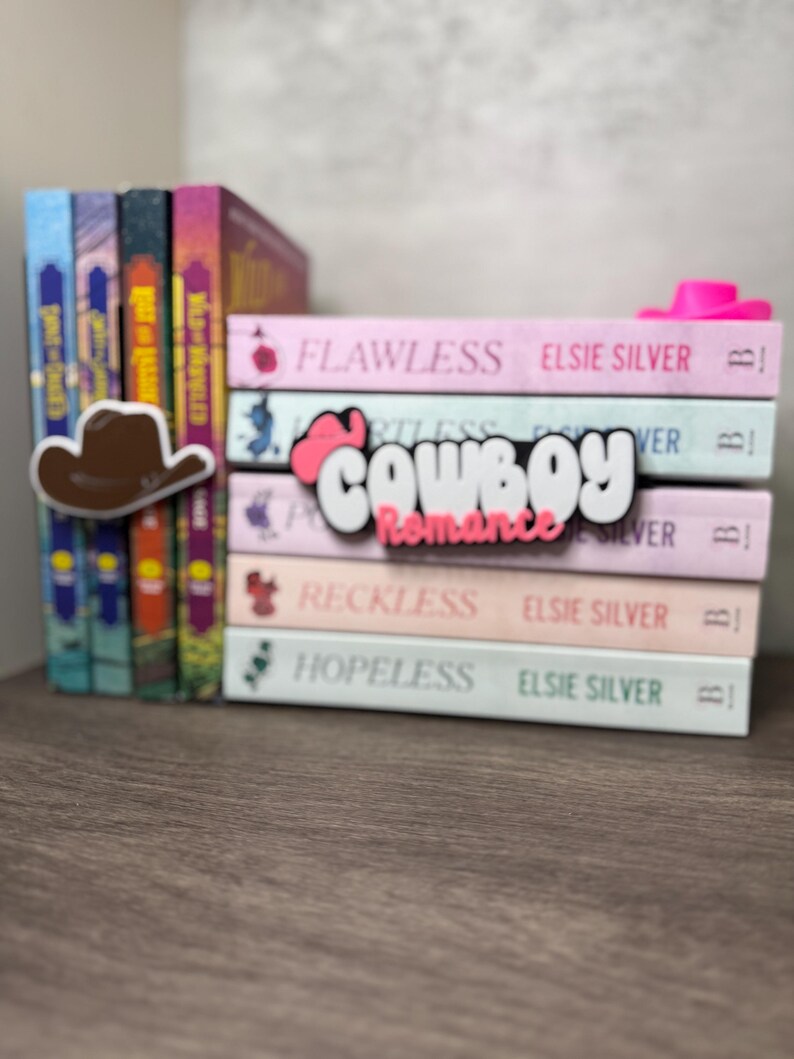 Puede incluir: Una pila de libros con cubiertas rosas, blancas y azules. Los libros se titulan "Flawless", "Reckless" y "Hopeless" de Elsie Silver. Un sombrero de vaquero marr&oacute;n y la palabra "Cowboy Romance" son visibles.