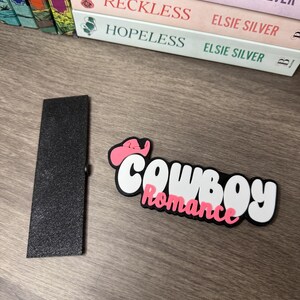 Puede incluir: Un sujetap&aacute;ginas rectangular negro y un accesorio para libros "Cowboy Romance" blanco, negro y rosa. El accesorio presenta un sombrero de vaquero rosa y la palabra "Romance" en rosa.