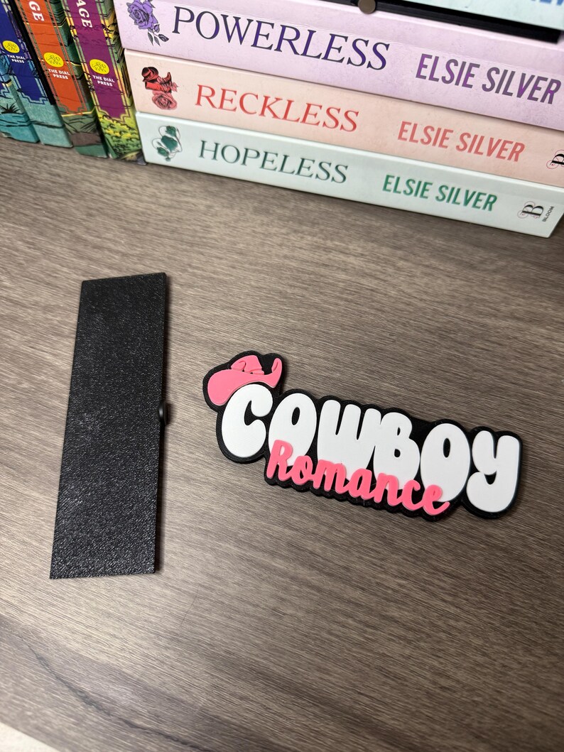 Puede incluir: Un sujetap&aacute;ginas rectangular negro y un accesorio para libros "Cowboy Romance" blanco, negro y rosa. El accesorio presenta un sombrero de vaquero rosa y la palabra "Romance" en rosa.