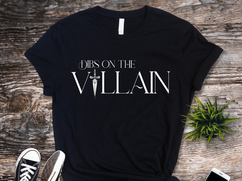 May include: Black t-shirt with the text "Dibs on the Villain" in white, featuring a sword graphic. The shirt is displayed on a wooden surface, with a small green plant and a pair of black and white sneakers visible.
