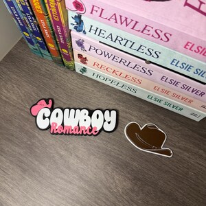 Puede incluir: Un juego de pegatinas decorativas con las palabras "Cowboy Romance" en una fuente juguetona, con una combinaci&oacute;n de colores rosa y blanco. Tambi&eacute;n se incluye una pegatina de sombrero de vaquero marr&oacute;n. Libros con t&iacute;tulos como "Flawless" y "Heartless" son visibles en el fondo.
