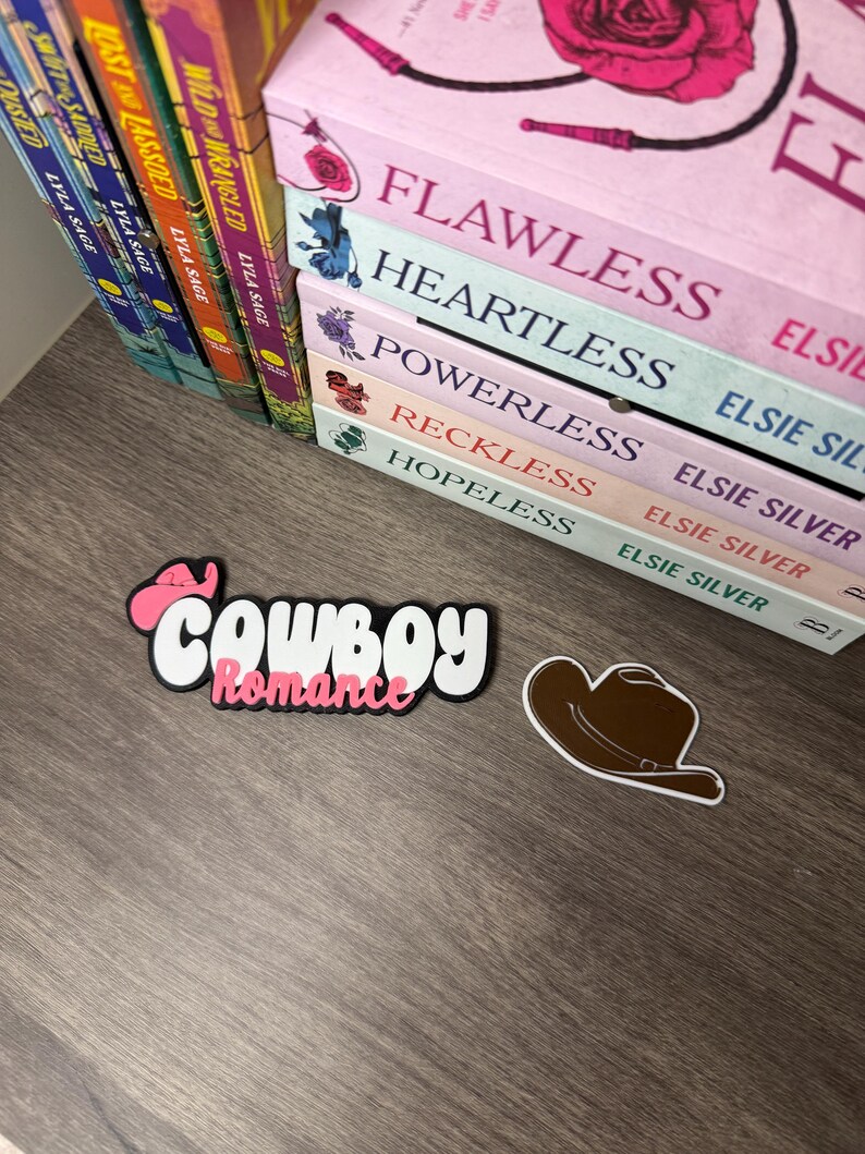 Puede incluir: Un juego de pegatinas decorativas con las palabras "Cowboy Romance" en una fuente juguetona, con una combinaci&oacute;n de colores rosa y blanco. Tambi&eacute;n se incluye una pegatina de sombrero de vaquero marr&oacute;n. Libros con t&iacute;tulos como "Flawless" y "Heartless" son visibles en el fondo.