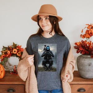 May include: Dark gray long-sleeve shirt featuring a graphic of a hooded figure with a blue skull face and a black cat. The figure is seated in a forest setting. The shirt is worn by a person wearing a brown hat and a tan cardigan.