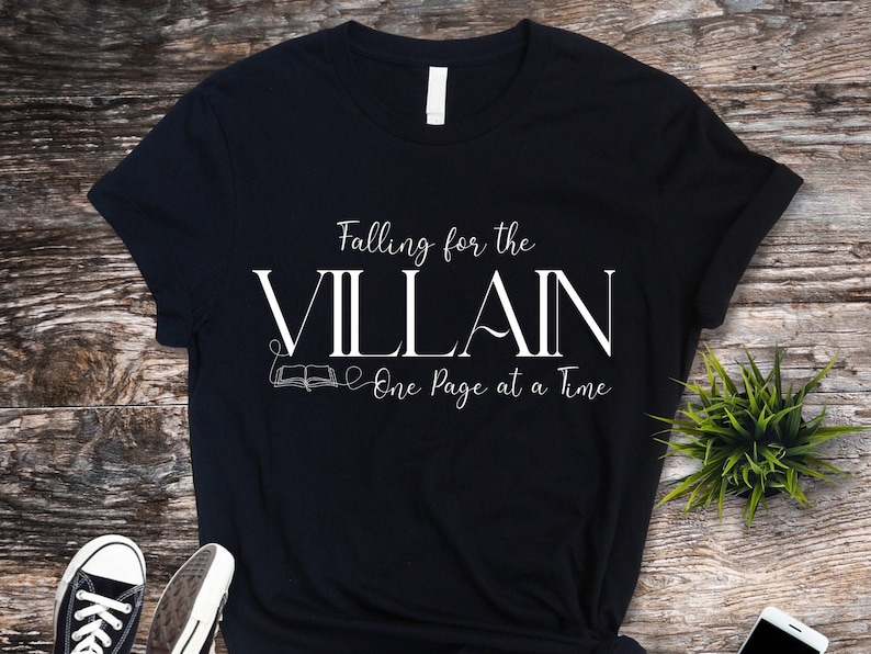 K&ouml;nnte beinhalten: Schwarzes T-Shirt mit dem wei&szlig;en Text "Falling for the VILLAIN One Page at a Time". Das Design beinhaltet eine Grafik eines offenen Buches. Das Shirt wird auf einer Holzoberfl&auml;che mit einer kleinen Pflanze und einem Smartphone pr&auml;sentiert.