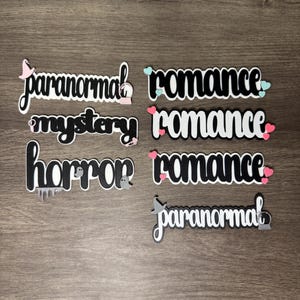 Puede incluir: Palabras troqueladas en blanco y negro, incluyendo "paranormal", "romance", "misterio" y "horror". Algunas palabras tienen elementos decorativos como corazones, fantasmas y sombreros de bruja. Los art&iacute;culos est&aacute;n dispuestos sobre una superficie de madera.